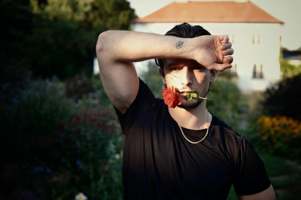 Young man with rose in mouth, arm with tattoo in garden setting.